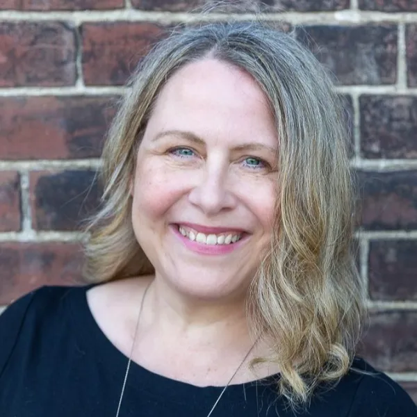 Portrait of a woman with light blond hair standing beside a brick wall.