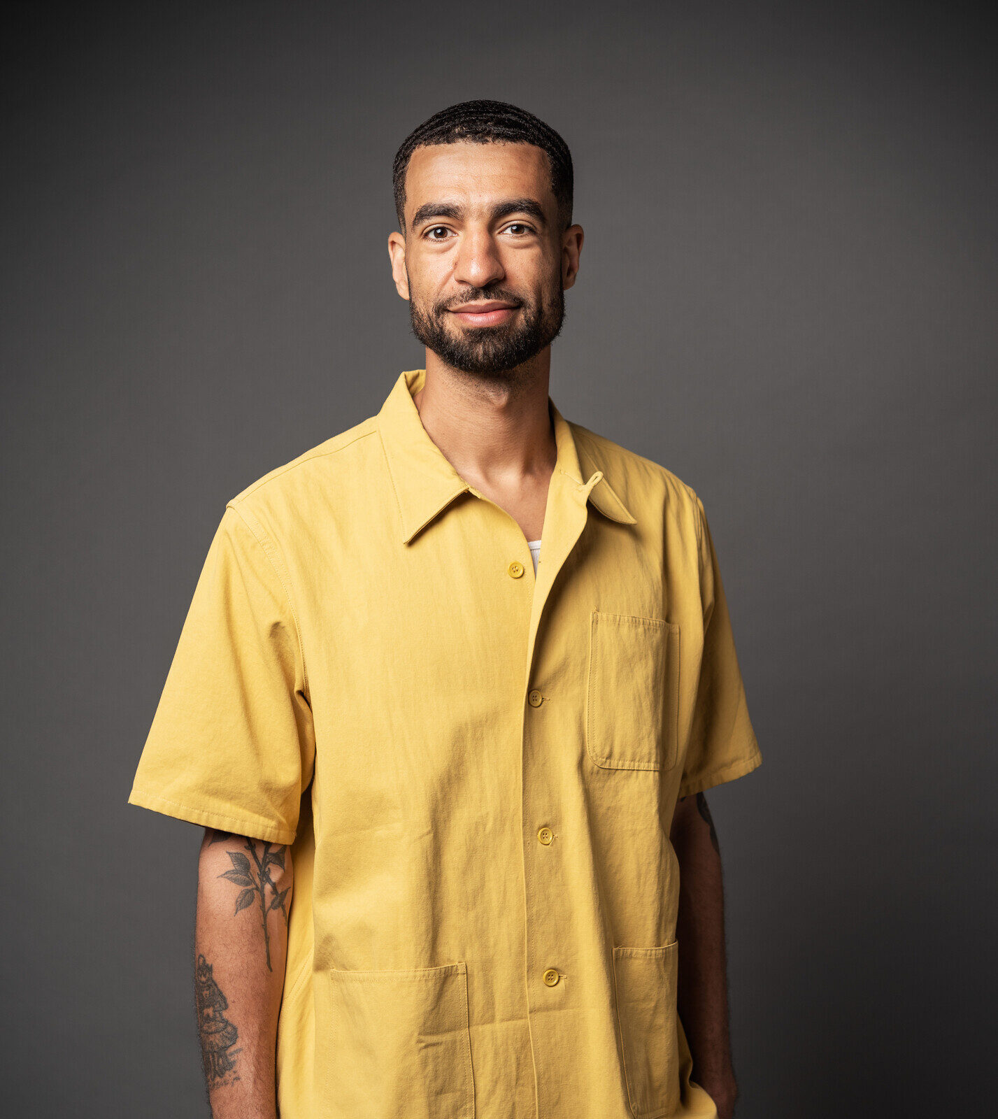 Portrait of a man wearing a loose yellow button-down shirt, with short black hair and a dark beard, posed in a studio.