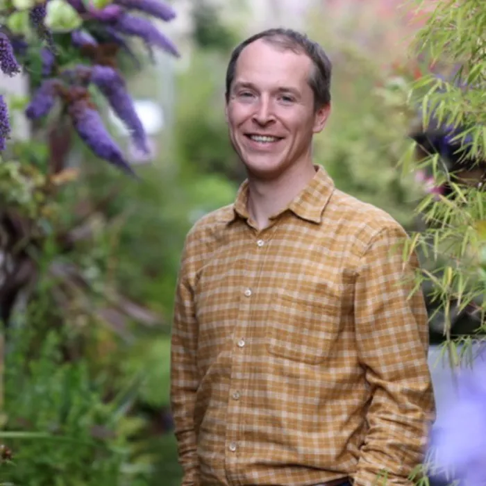 Smiling man with dark brown hair and yellow plaid button down shirt standing in a garden with purple flowers