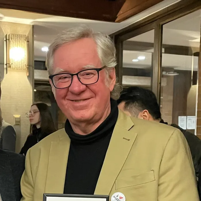 Smiling man with white hair, black turtleneck, and mustard-khaki blazer