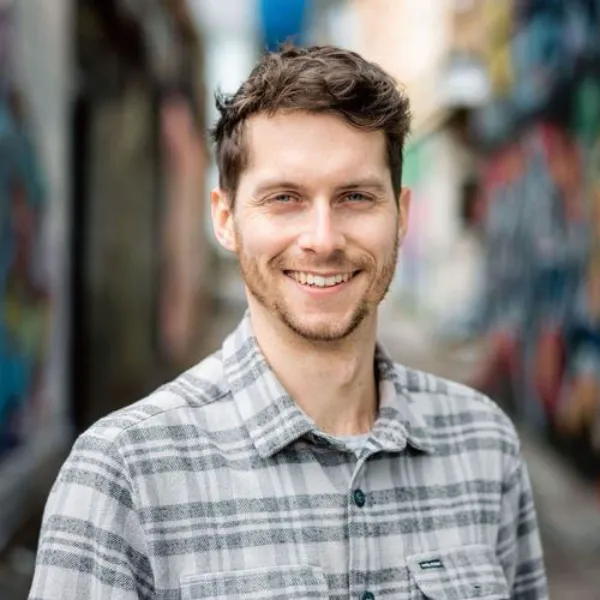 Portrait of a man with light brown hair and a beard, wearing a beige plaid button-down shirt, standing in a blurry alley background.